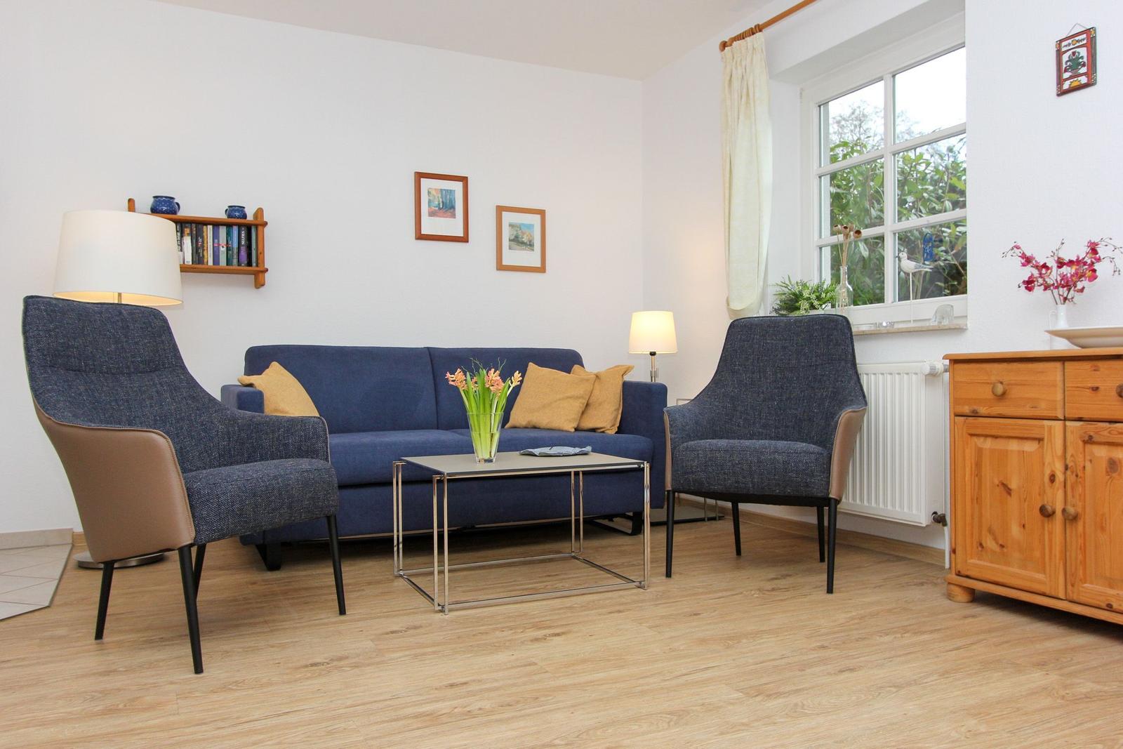Ein Wohnzimmer mit blauem Sofa, zwei Sesseln und Holzmöbeln. An der Wand hängen Bilder.