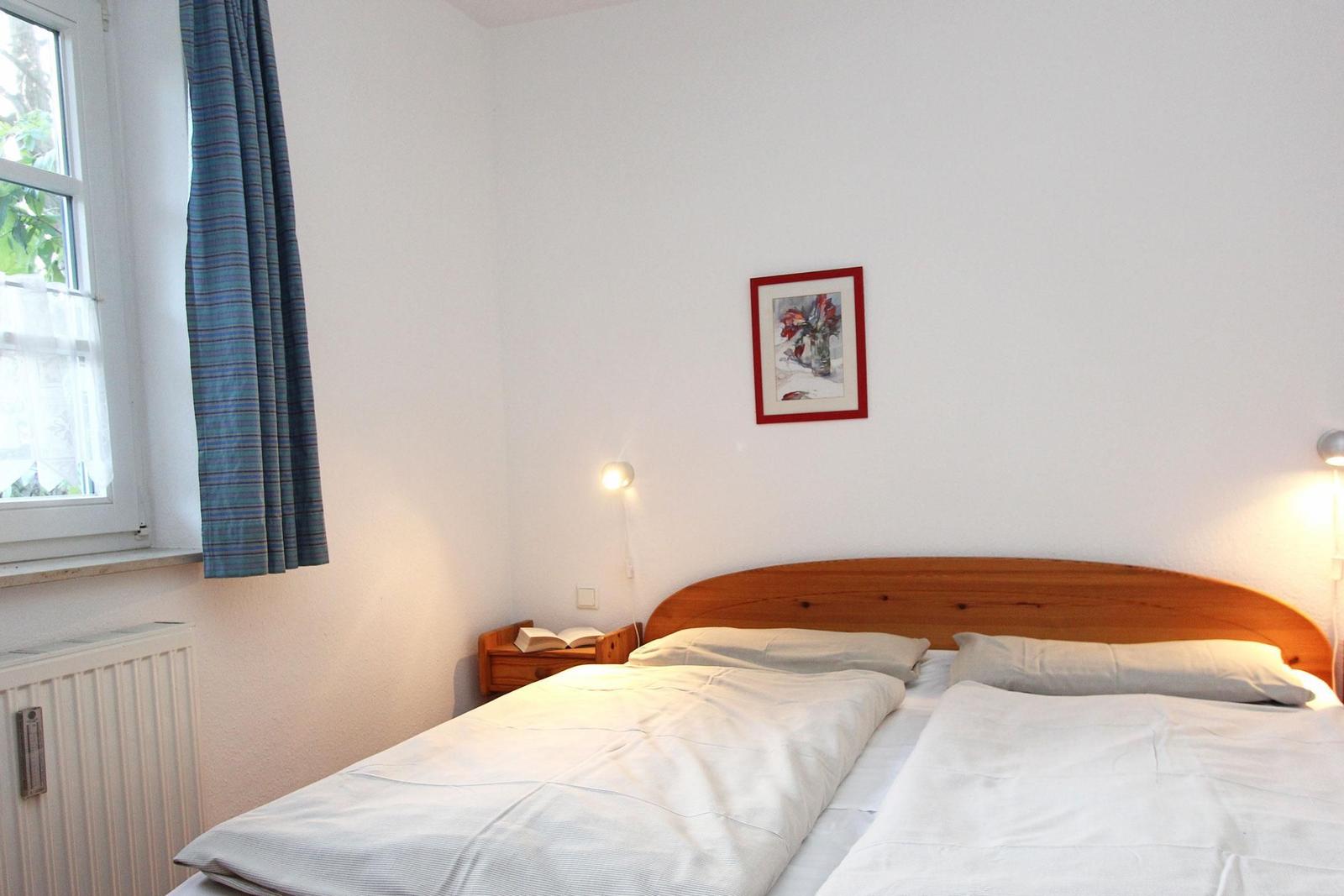 Ein Schlafzimmer mit Doppelbett, Holzkopfteil, Nachttisch und Fenster mit Vorhang.