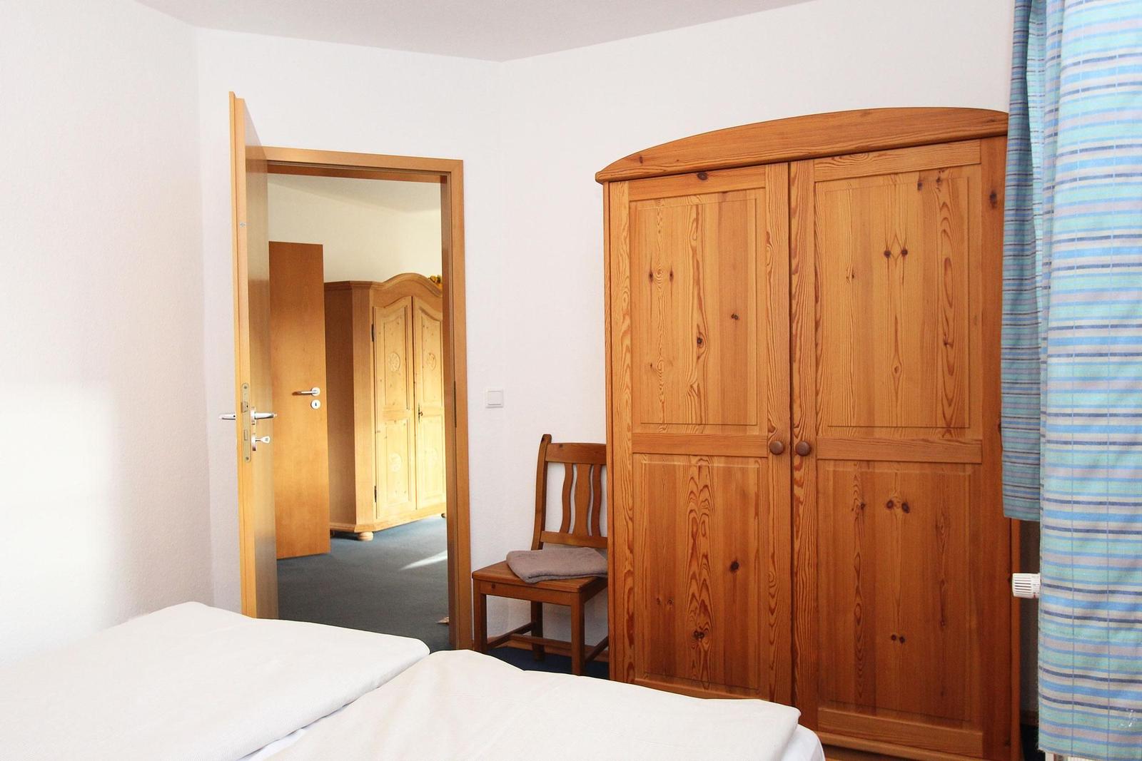 Schlafzimmer mit Holzschrank, Stuhl und Blick in angrenzenden Raum.