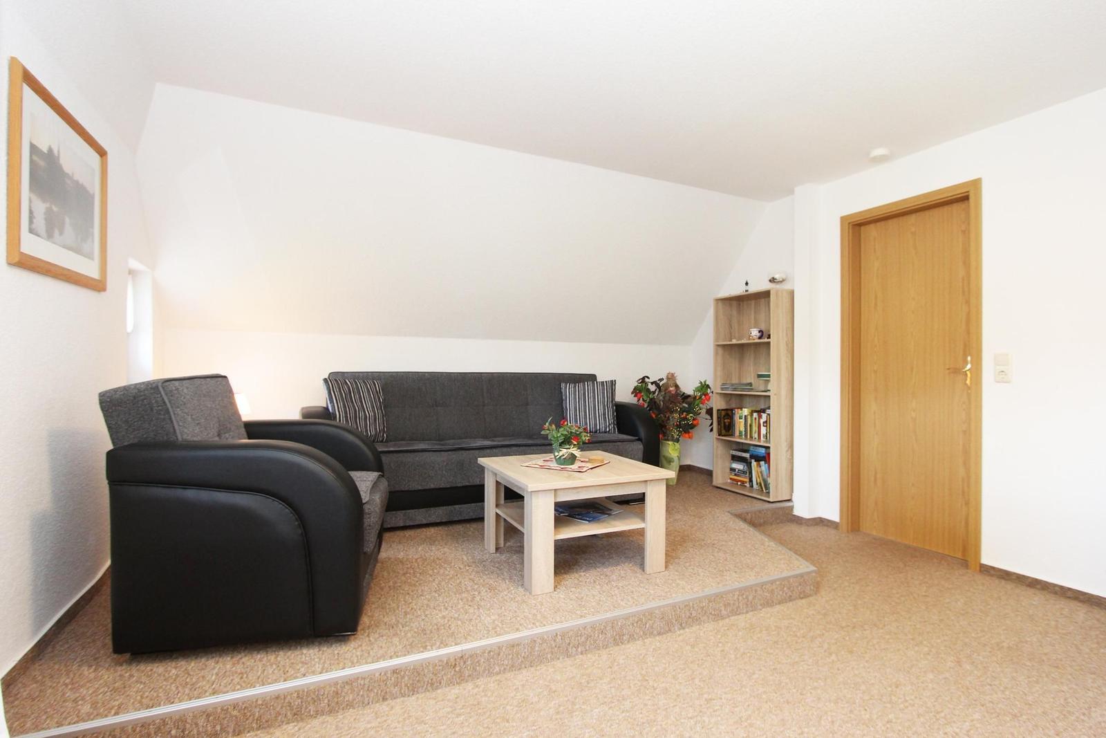 Wohnzimmer mit Sofa, Sessel, Tisch und Bücherregal. Holztür und Teppichboden.