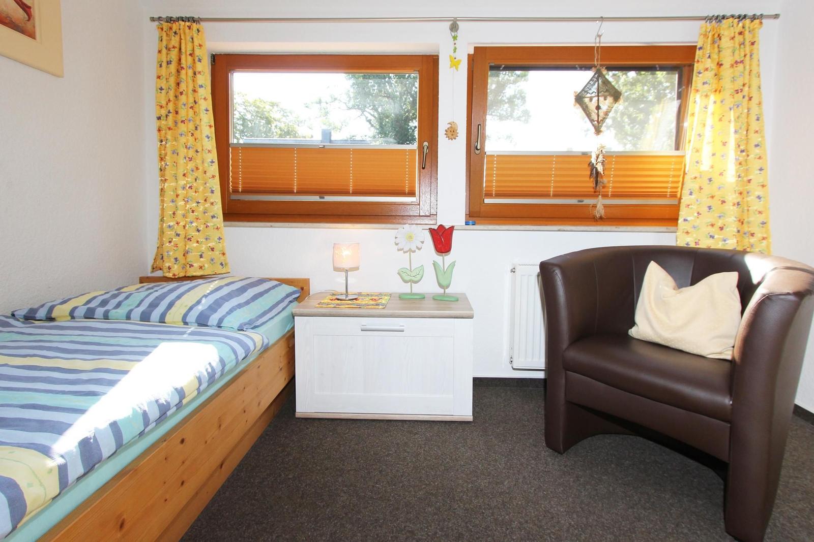Ein Zimmer mit Bett, Sessel und zwei Fenstern mit Jalousien und Vorhängen.