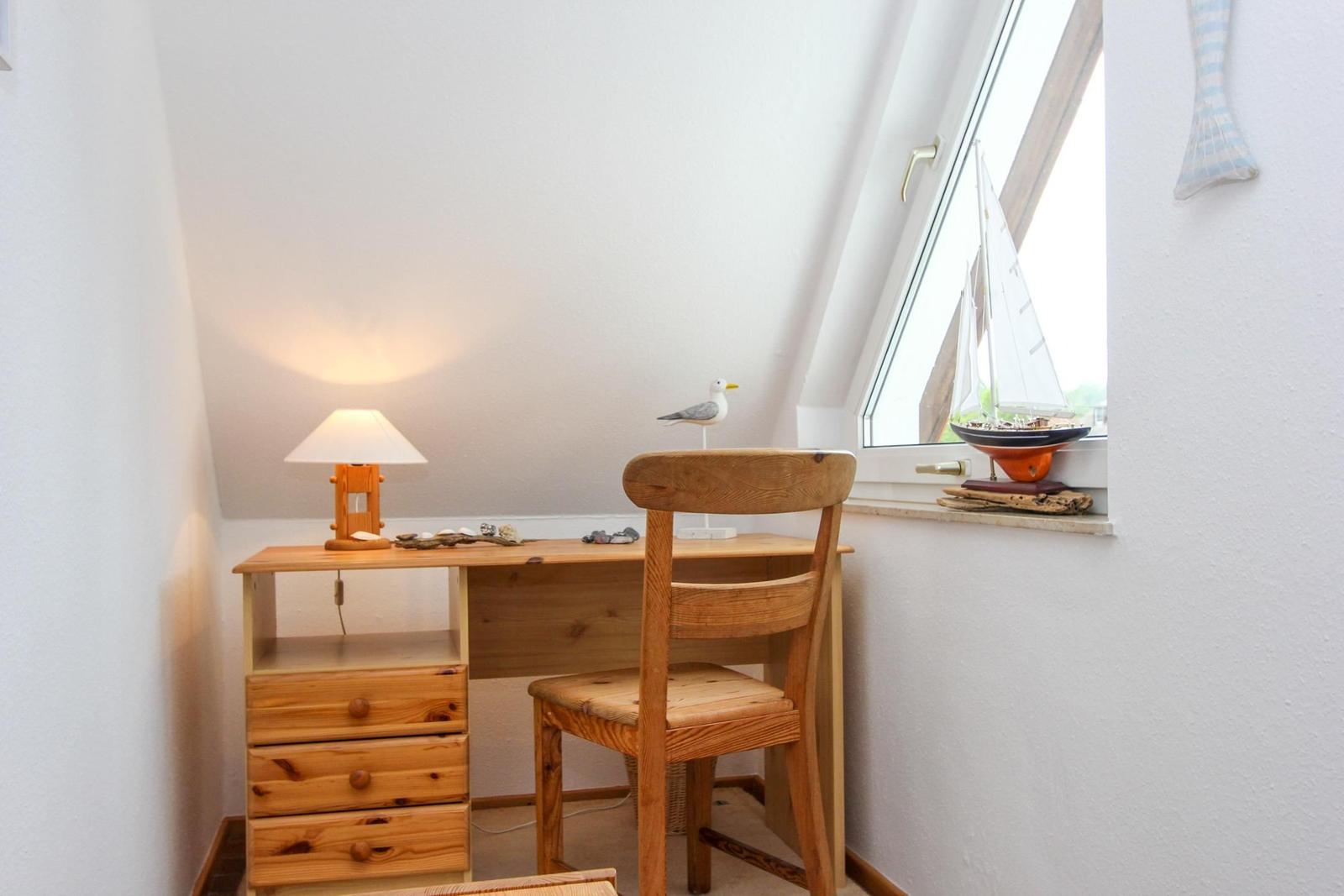 Schreibtisch mit Stuhl und Lampe unter Dachschräge. Modellboot und Vogel auf Fensterbank.