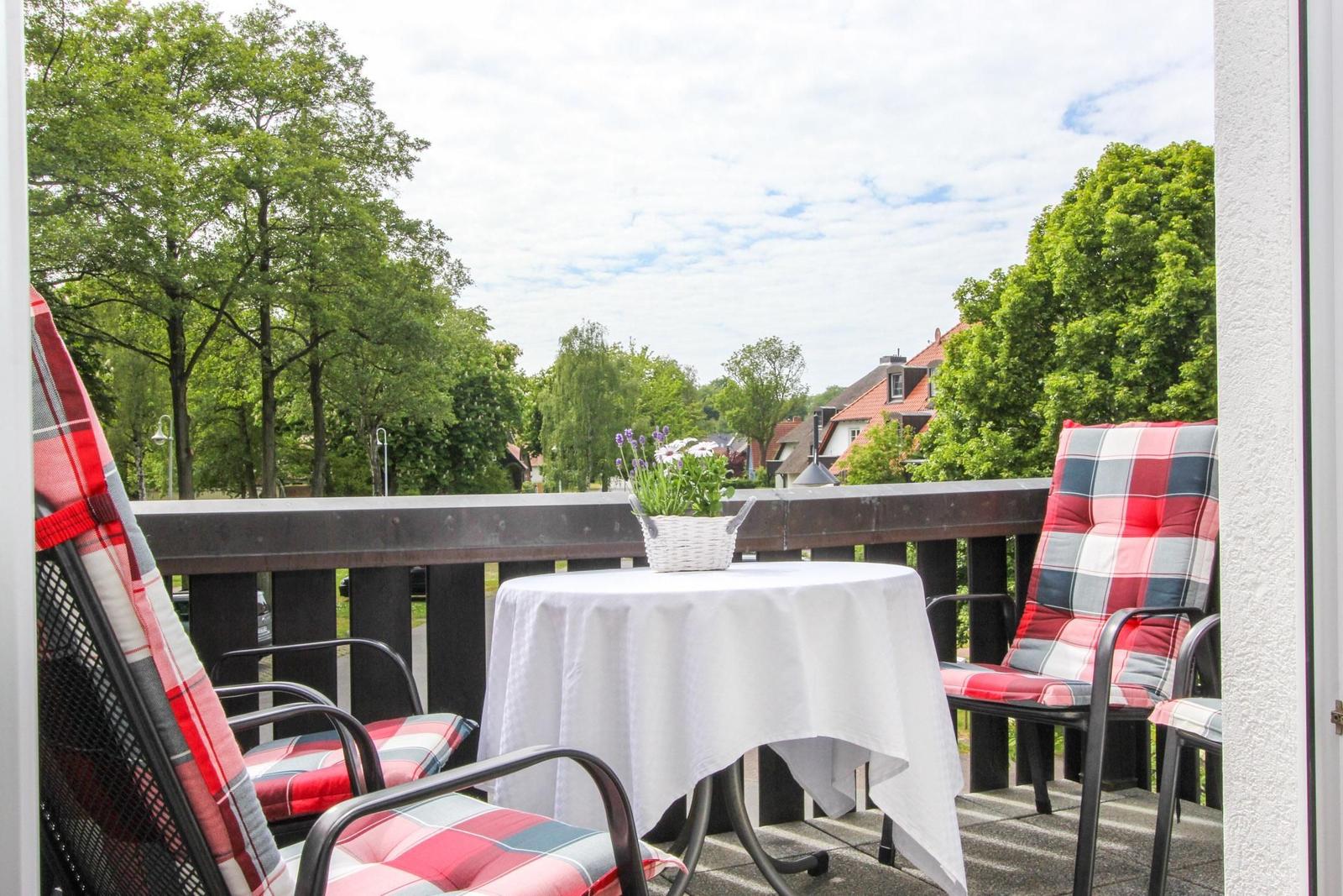 Terrasse mit Tisch, zwei Stühlen und Blumenkorb. Blick auf grüne Bäume und Häuser.
