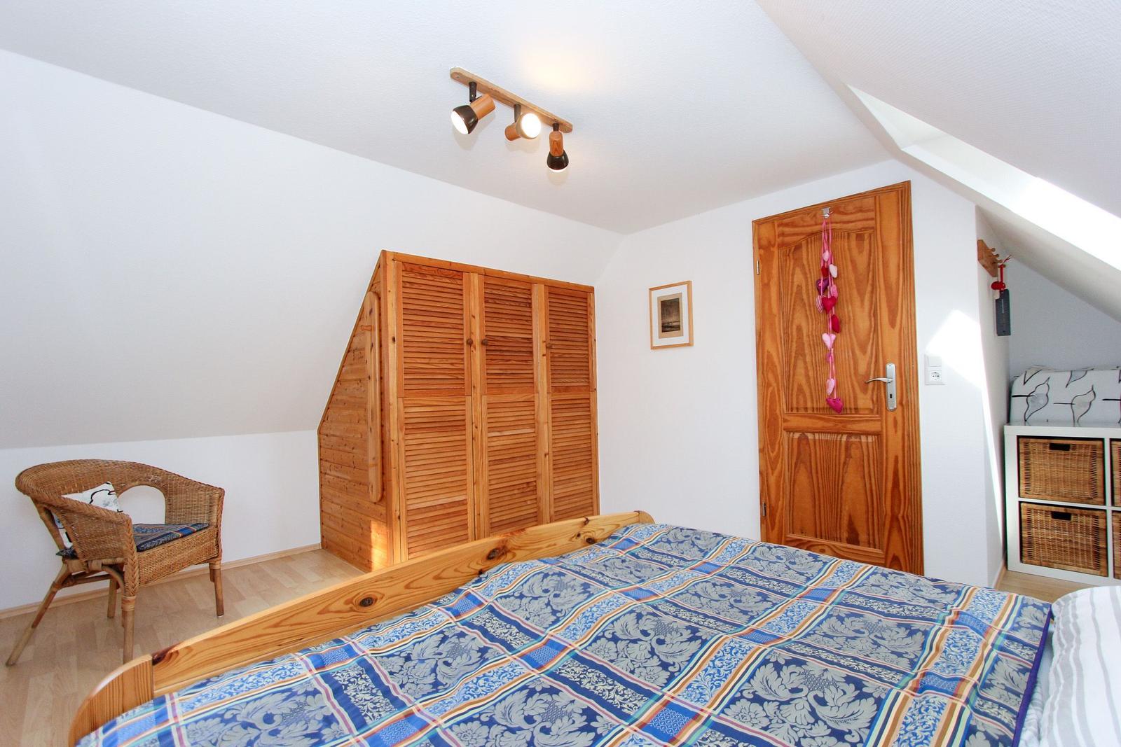 Schlafzimmer mit Holzbett, Kleiderschrank und Wickerstuhl unter Dachschrägen.