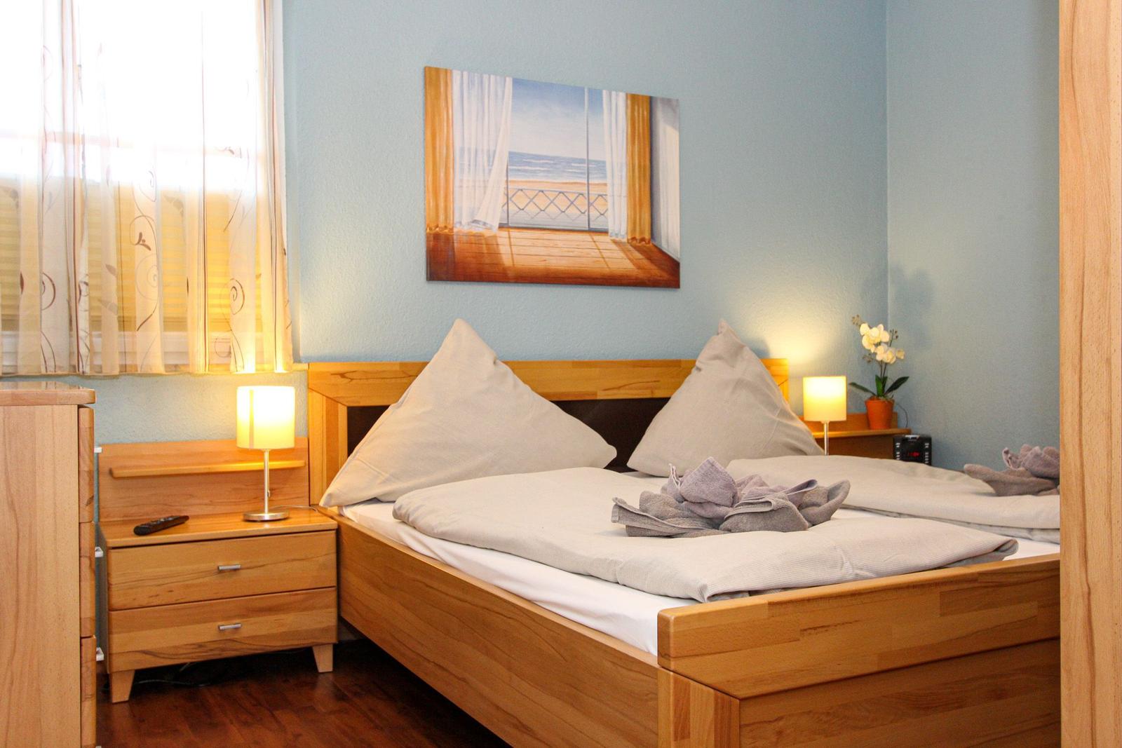 Schlafzimmer mit Holzbett, Nachttisch und Wandbild. Fenster mit Vorhängen.
