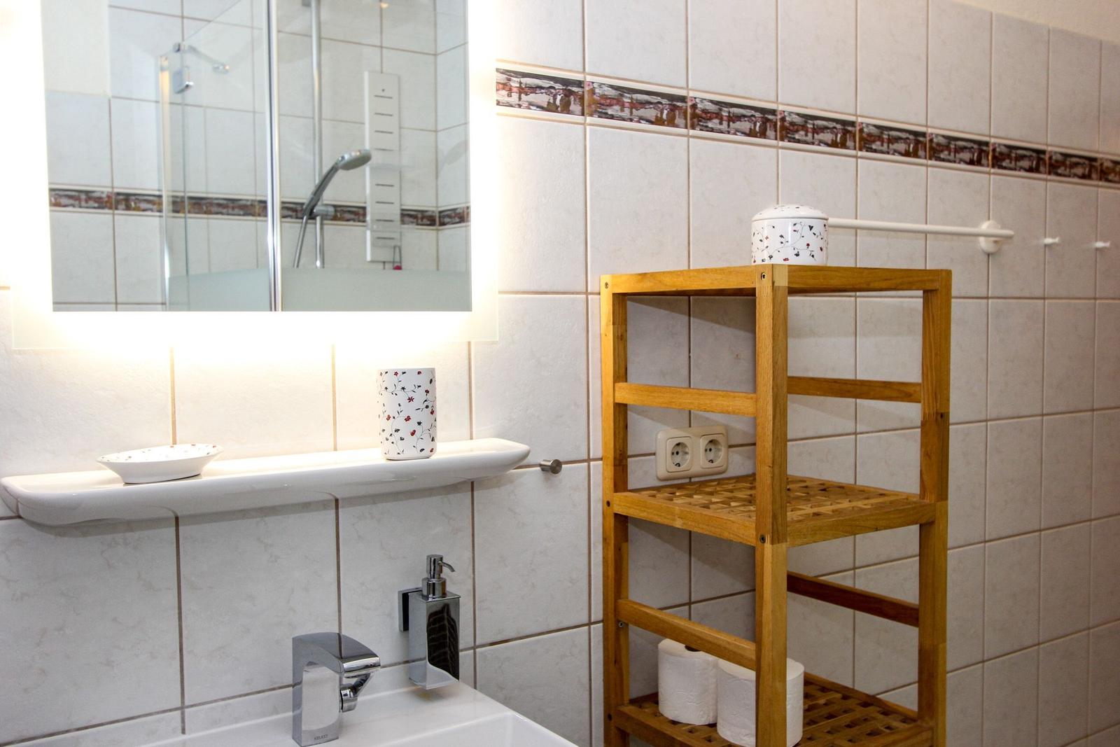 Badezimmer mit Waschbecken, Spiegel und Holzregal. Fliesenwand mit dekorativer Bordüre.