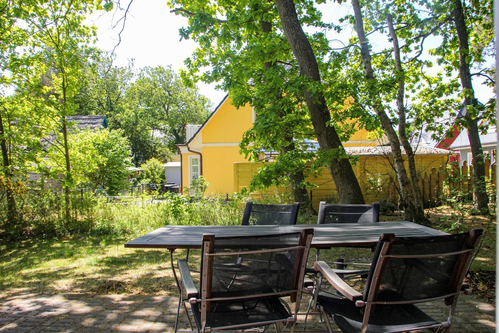 Hinter einem Tisch mit Stühlen steht ein gelbes Haus unter Bäumen.