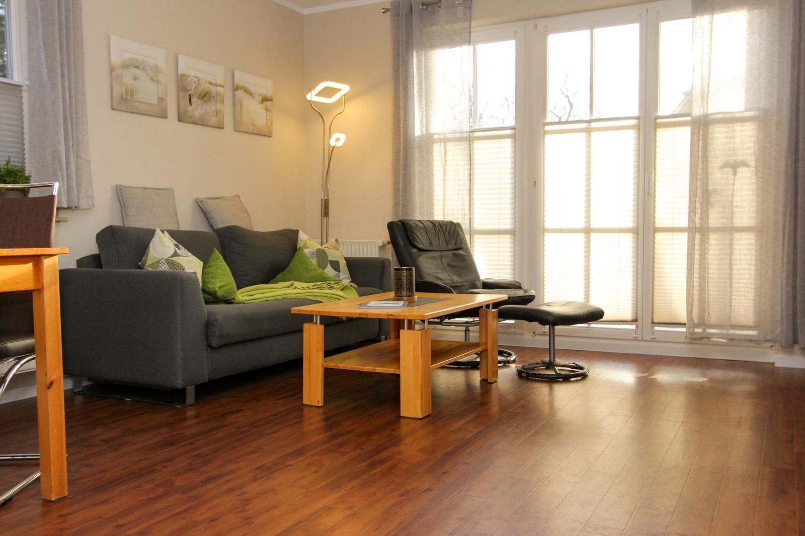 Wohnzimmer mit Sofa, Sessel, Tisch und Holzboden. Große Fenster mit Jalousien.