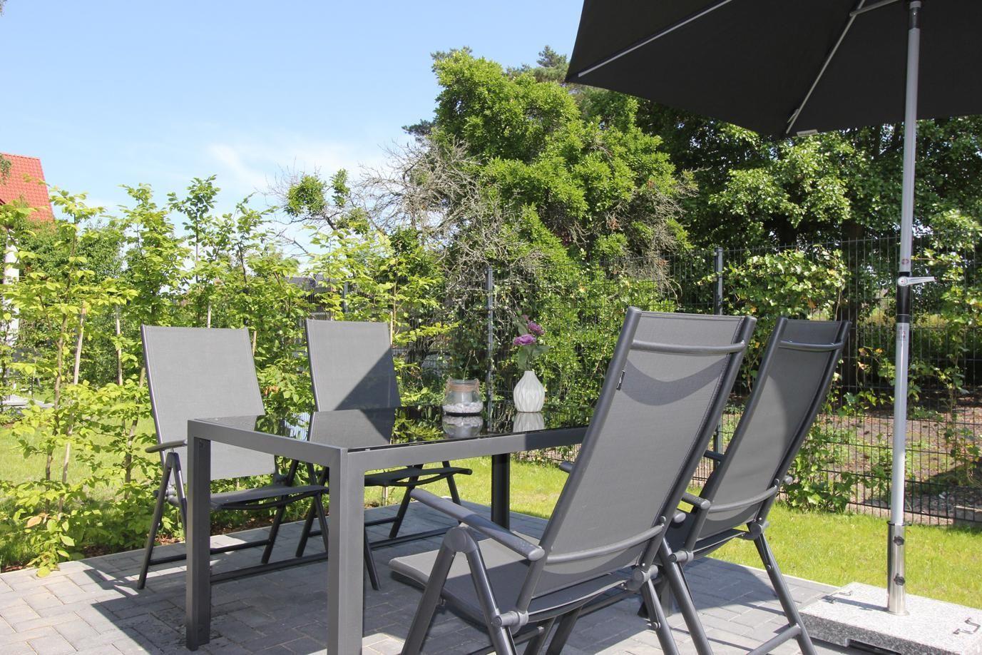 Terrasse mit Tisch, vier Stühlen und Sonnenschirm. Grün hinter dem Zaun.