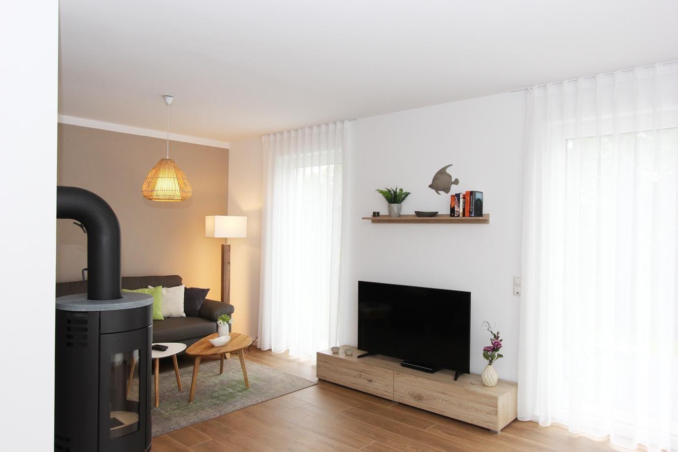 Wohnzimmer mit Sofa, Fernseher und Kaminofen. Fenster mit weißen Vorhängen.