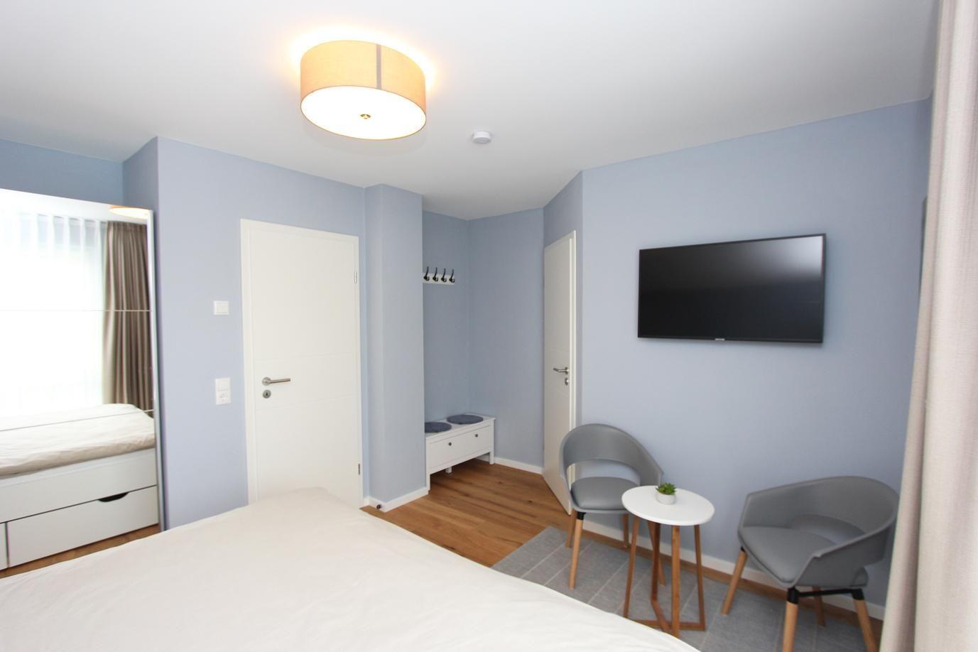 Schlafzimmer mit Bett, Fernseher und zwei Stühlen. Wandfarbe hellblau, Holzboden.