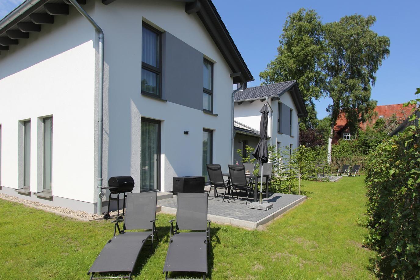 Moderne Villa mit Terrasse, Gartenmöbeln und Grillplatz. Grünflächen und Bäume umgeben das Haus.
