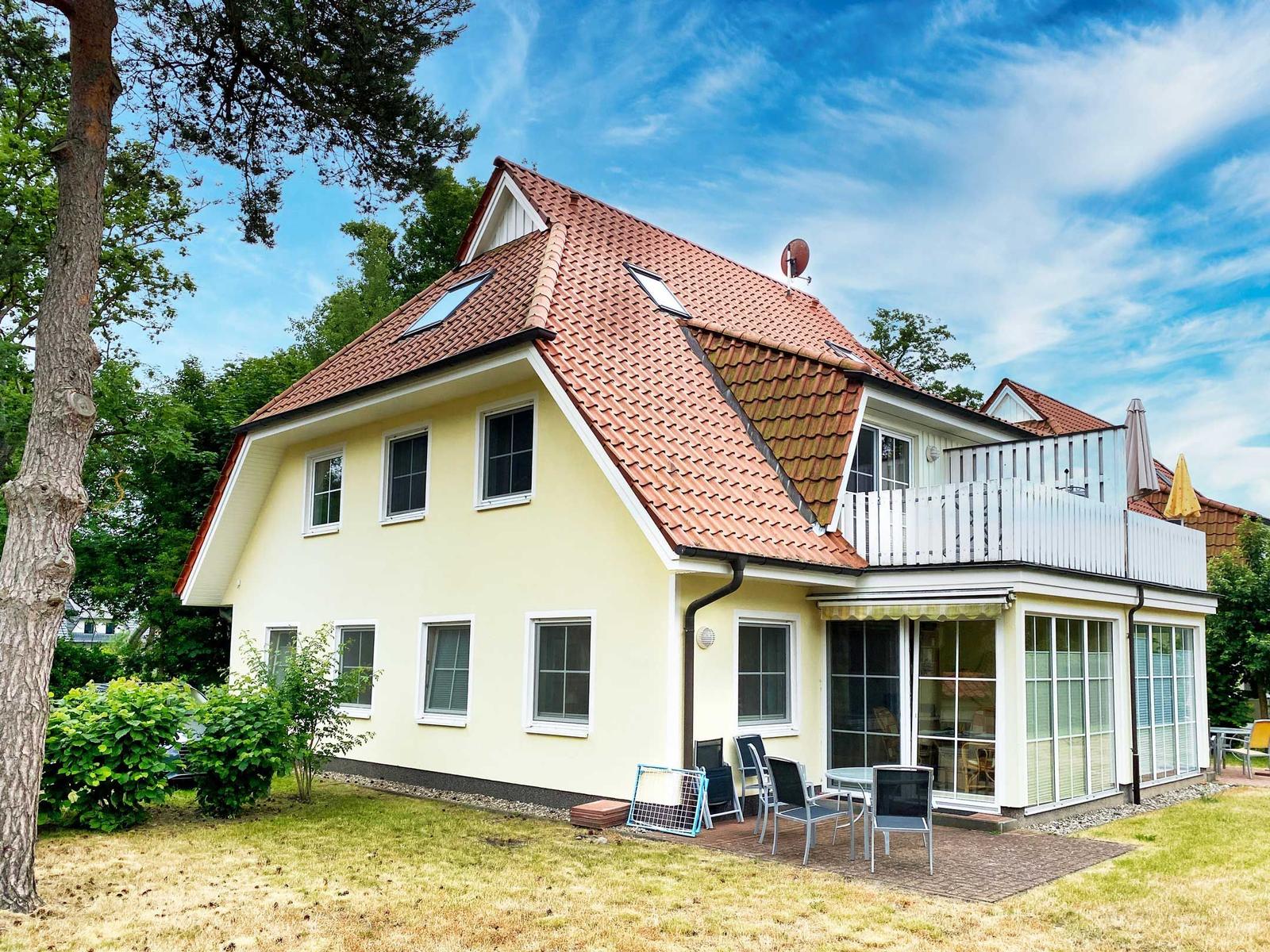 Ein zweistöckiges Haus mit rotem Dach und Terrasse. Garten und Sitzgelegenheiten sind vorhanden.