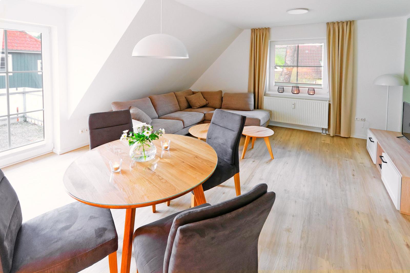 Wohn- und Essbereich mit Sofa, Tisch und Stühlen. Holzboden und große Fenster.