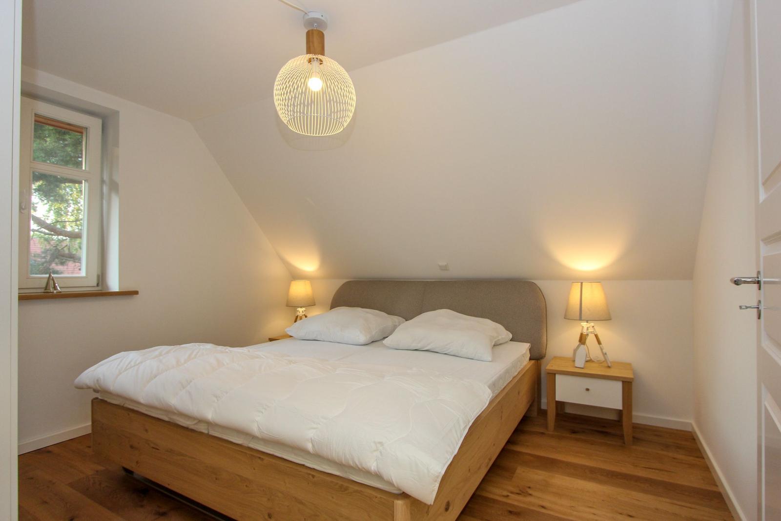 Schlafzimmer mit Doppelbett, Nachttisch und Fenster. Holzboden und Deckenleuchte.