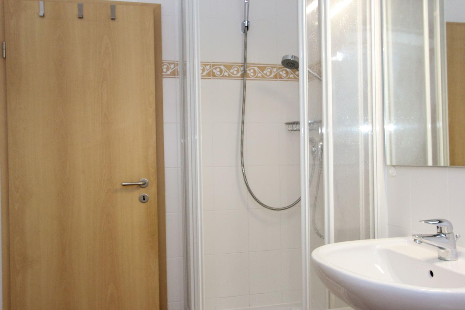 Badezimmer mit Dusche, Waschbecken und Spiegel. Holztür und weiße Fliesen.