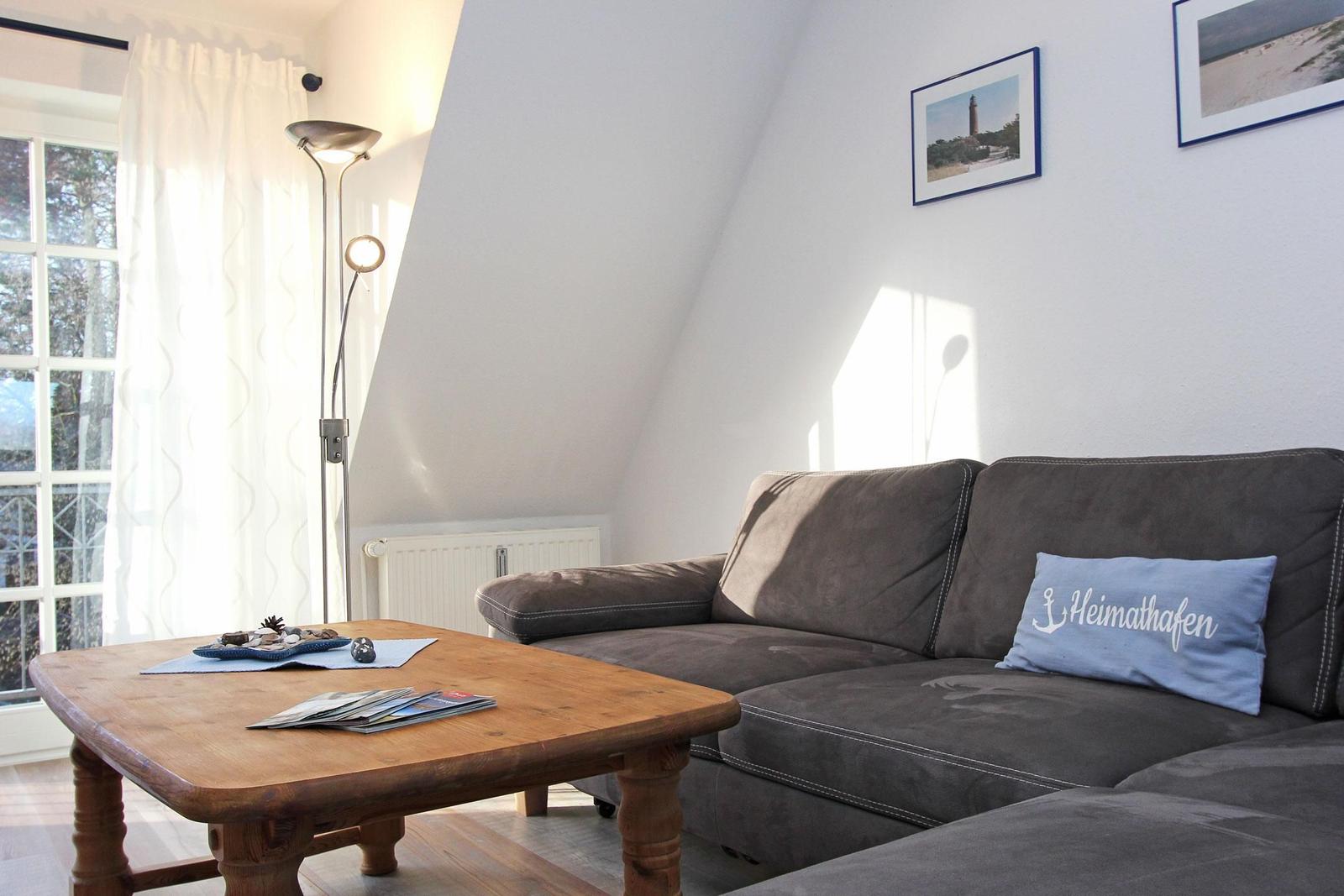 Ein Wohnzimmer mit grauem Sofa, Holztisch und Fenster mit weißen Vorhängen.