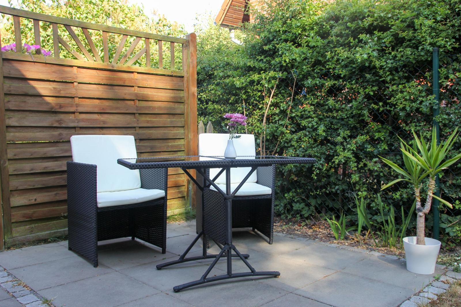 Terrasse mit zwei Stühlen und Tisch, umgeben von Hecke und Holzzaun.