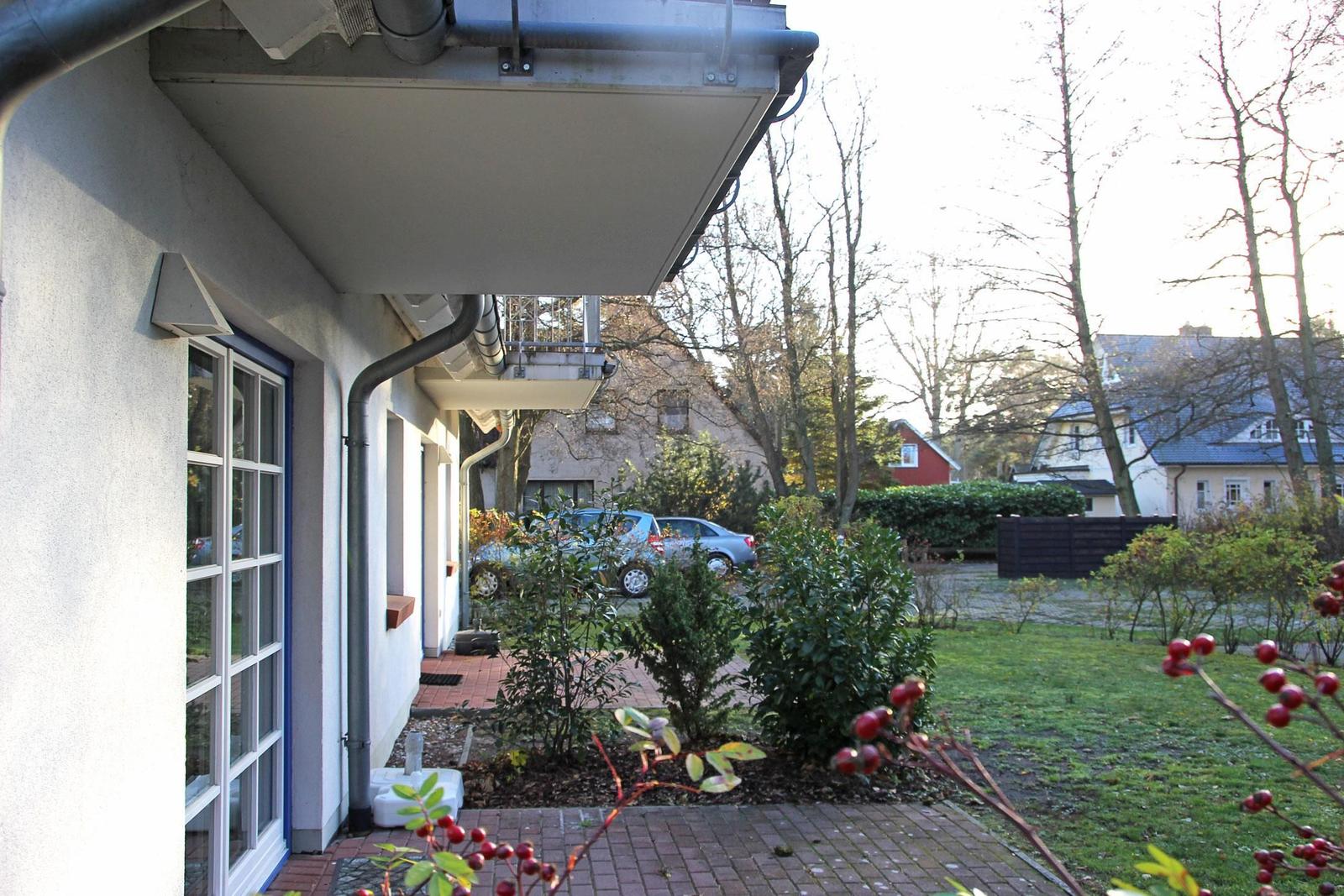 Vor dem Haus mit Balkon, Garten und Parkplatz. Bäume und Sträucher im Hintergrund.