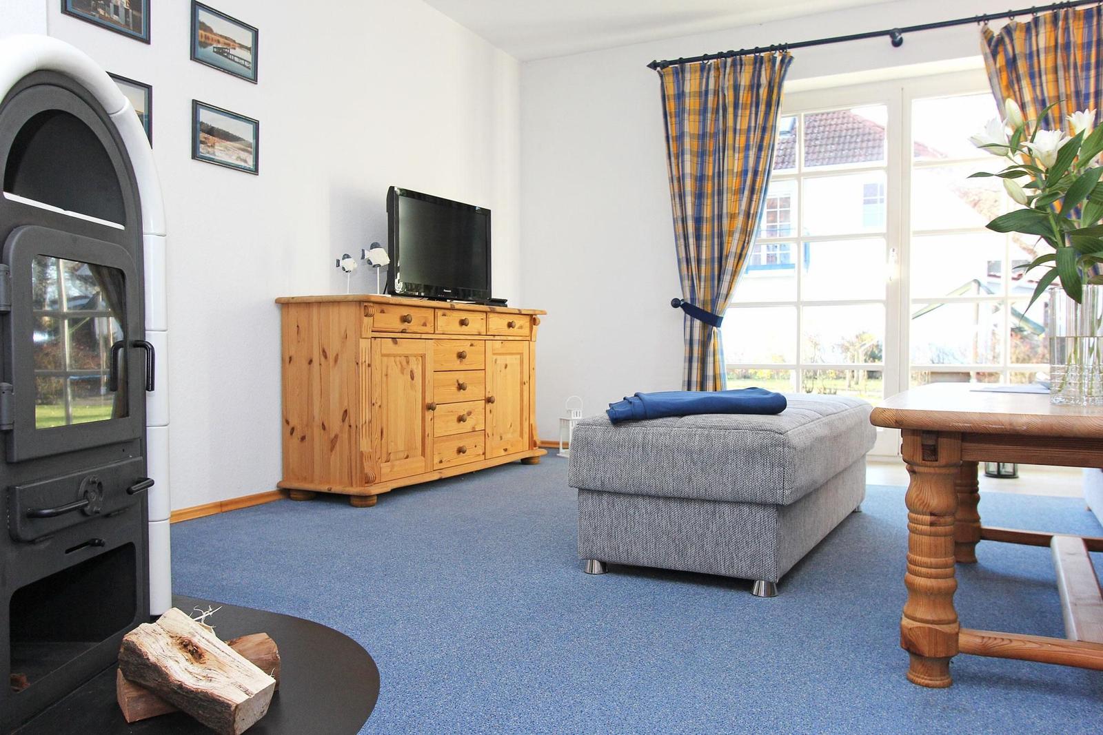 Wohnzimmer mit Kamin, Fernseher, Sitzbank und Tisch. Blaue Teppichboden und Fenster mit Vorhängen.
