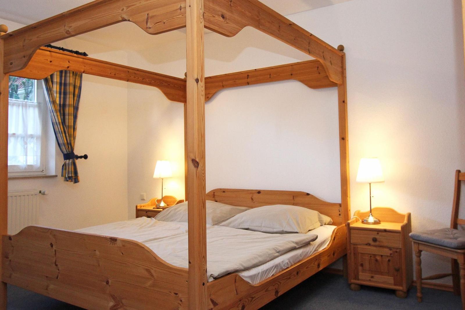 Schlafzimmer mit Holzbett, Nachttischen und Stuhl. Fenster mit Vorhängen.