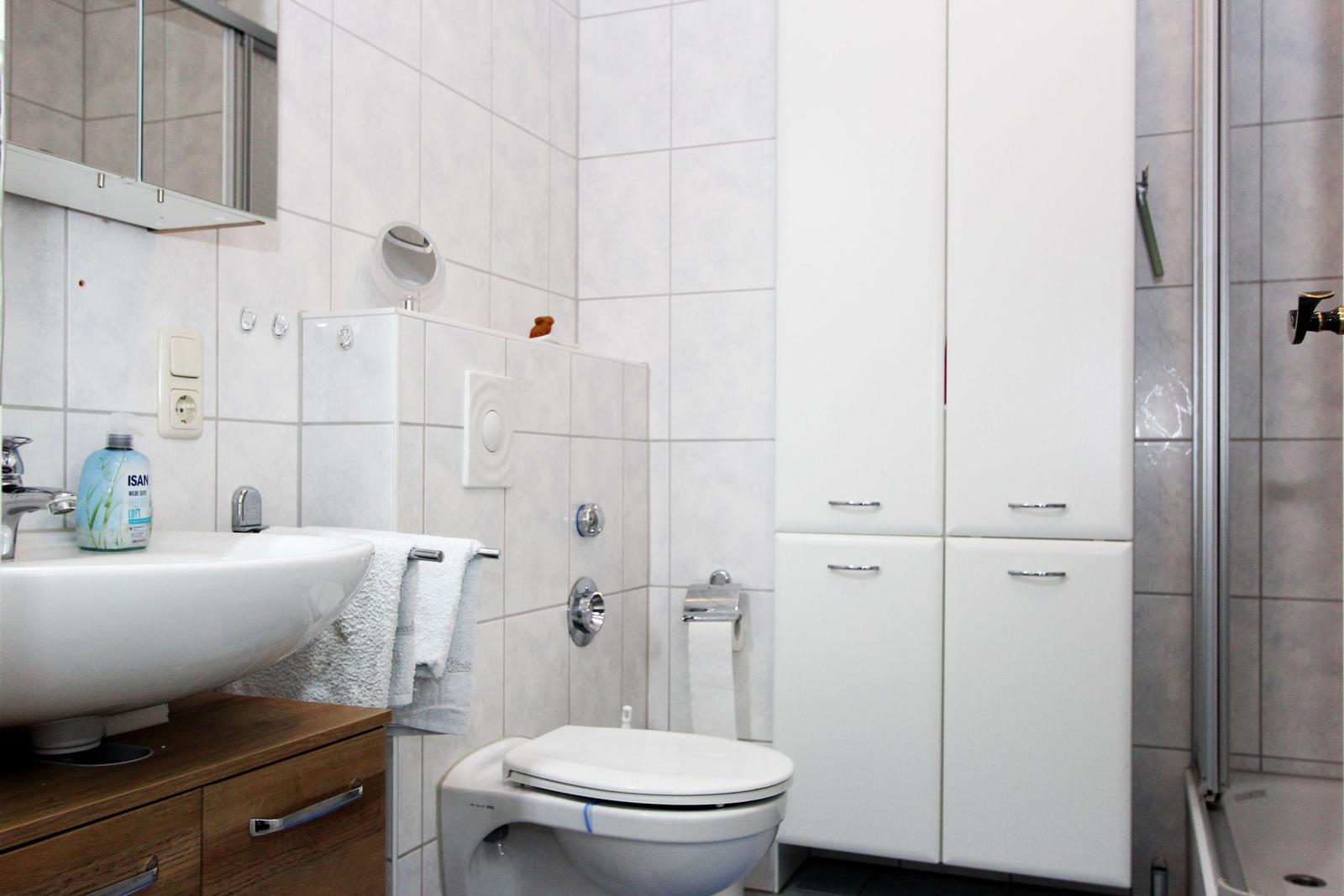 Badezimmer mit Waschbecken, Toilette und Dusche. Weiße Fliesen und Schränke.