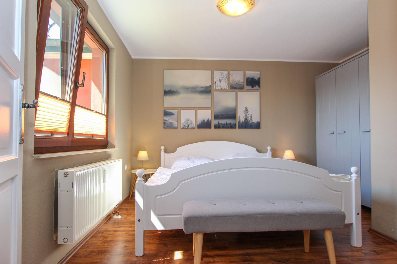 Schlafzimmer mit weißem Bett, grauer Sitzbank, Wandbildern und Fenster mit Jalousie.