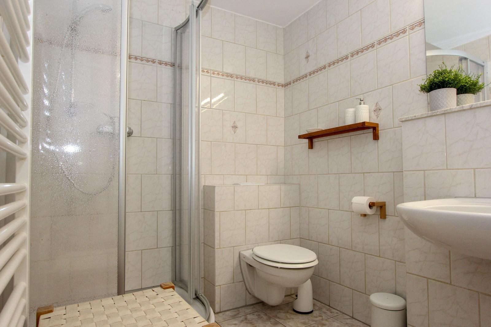 Badezimmer mit Dusche, Toilette und Waschbecken. Weiße Fliesen und Holzregal.