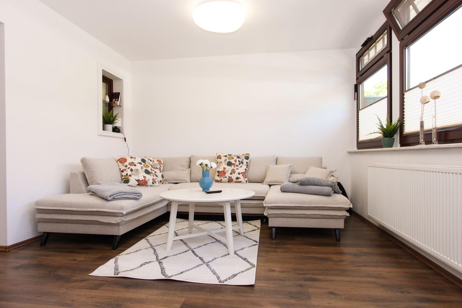 Wohnzimmer mit Ecksofa, runden Tisch und Holzboden. Fenster mit Jalousien und Pflanzen.