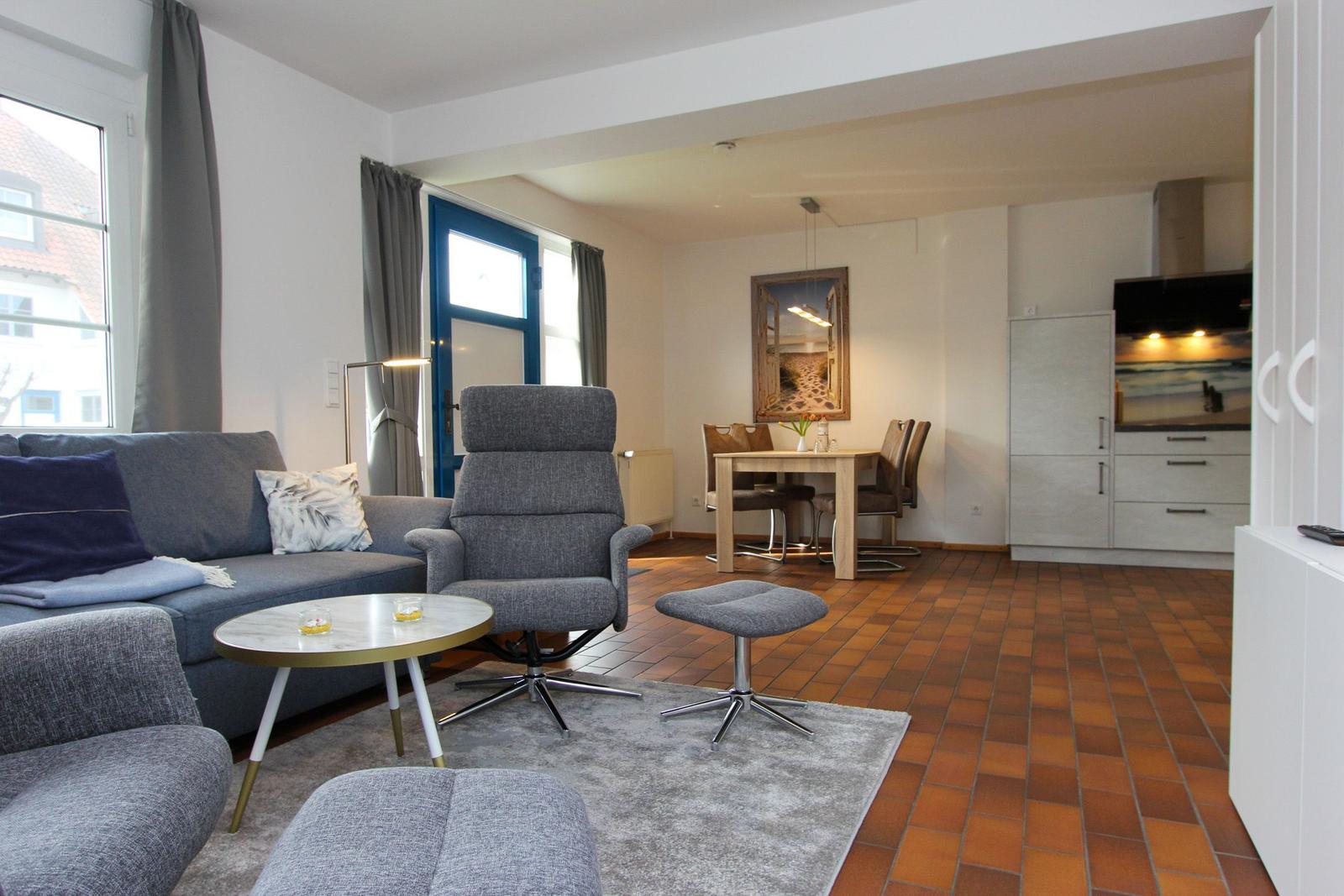 Wohnzimmer mit Sofa, Sessel und Esstisch. Küche im Hintergrund. Terrakottaboden.