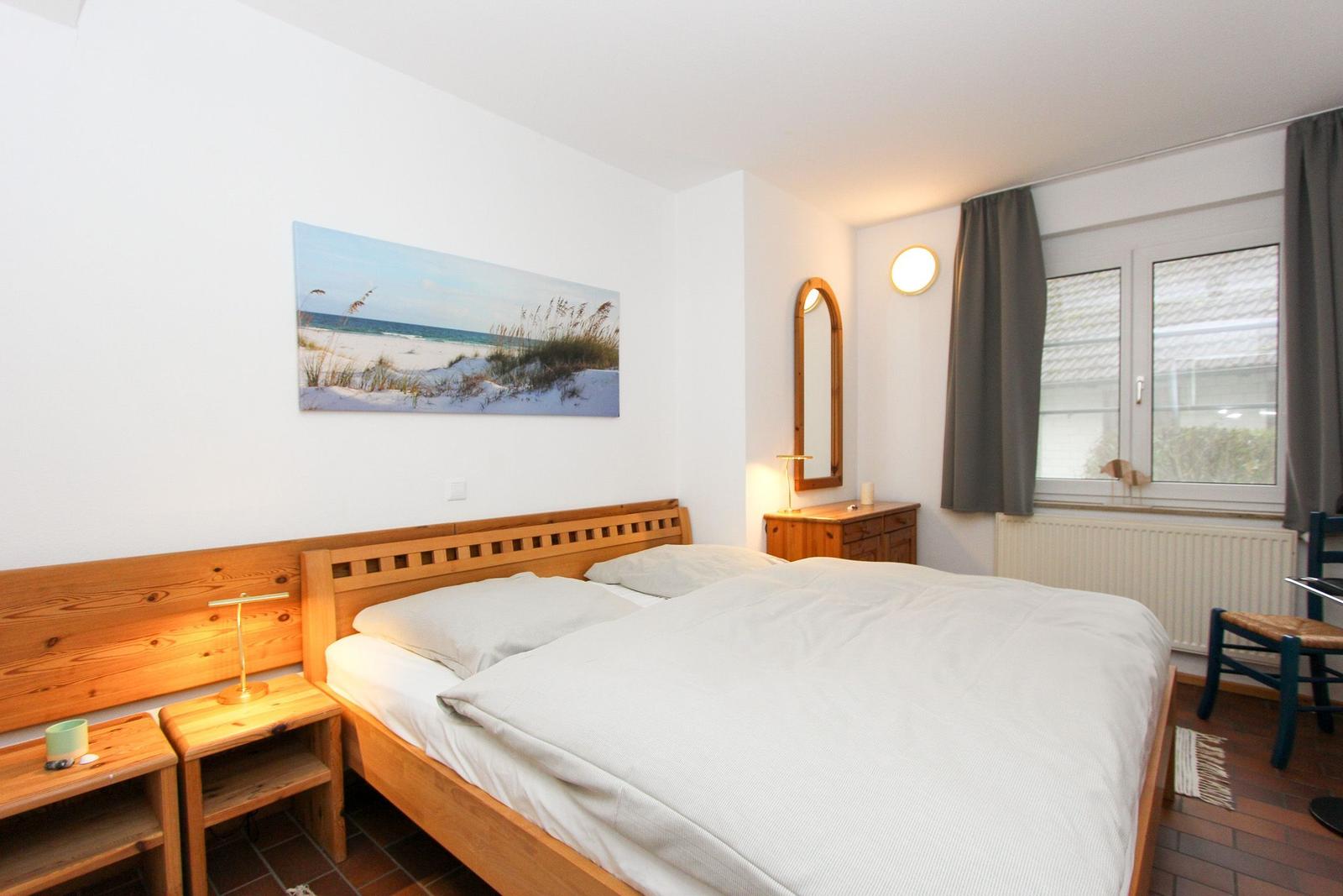 Ein Schlafzimmer mit Holzbett, Nachttisch und Fenster. An der Wand hängt ein Strandbild.