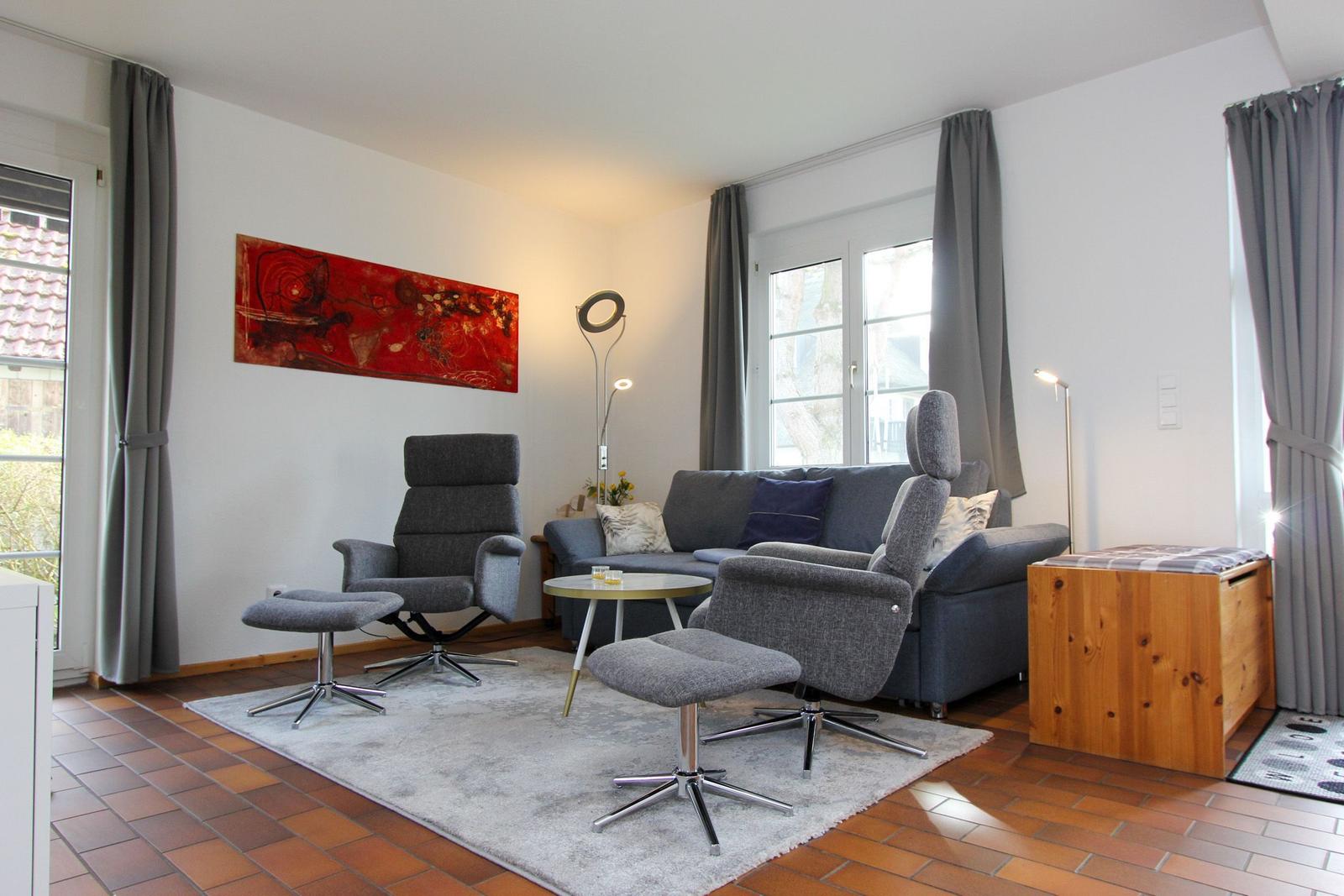 Wohnzimmer mit Sofa, zwei Sesseln, Tisch und Wandbild. Holzfußboden und Fenster mit Vorhängen.