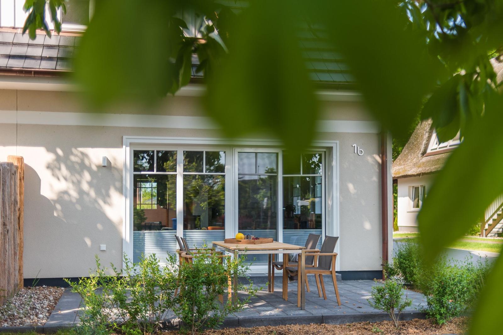 Haus mit Terrasse, Tisch und Stühlen. Grüne Pflanzen und Bäume umgeben das Gebäude.