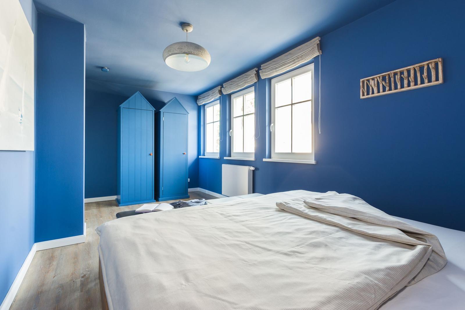 Schlafzimmer mit blauen Wänden, zwei Fenstern und einem Bett mit weißer Bettwäsche.
