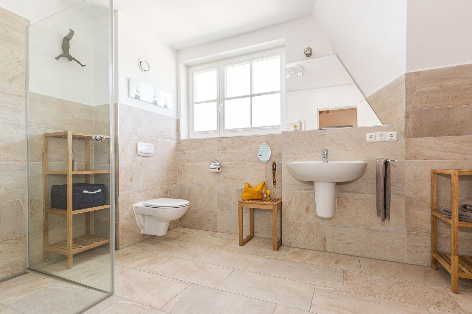 Badezimmer mit Dusche, Waschbecken und Toilette. Holzregale und Fenster sichtbar.