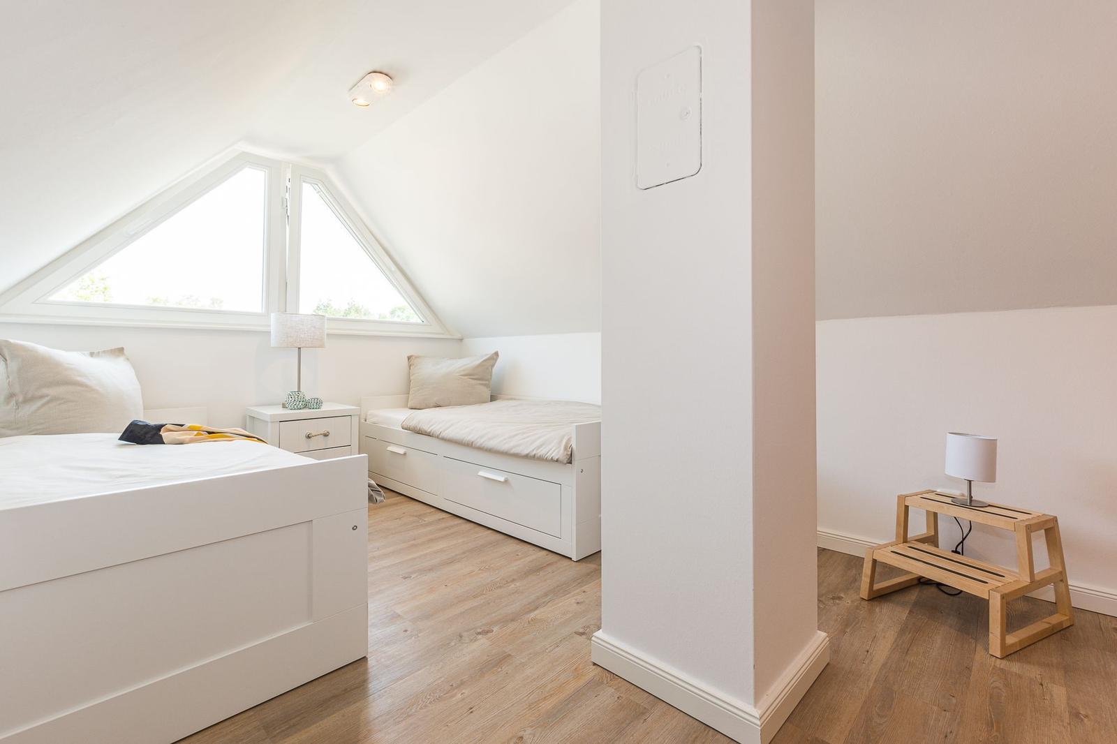 Zimmer mit Dachschrägen, zwei Betten und Holzboden. Fenster mit Blick nach draußen.