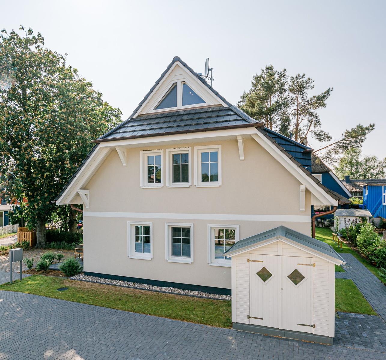 Ein zweistöckiges Haus mit weißem Anstrich und grauem Dach. Vor dem Haus steht ein kleiner weißer Schuppen.