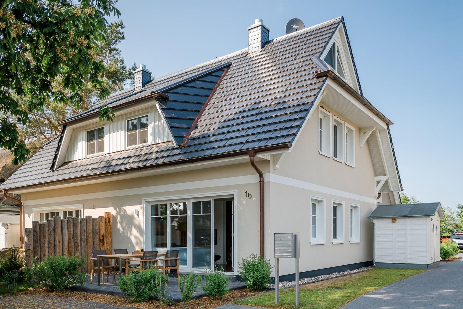 Ein Haus mit Dachgauben, Terrasse und Garten. Hausnummer 1b. Satellitenschüssel auf dem Dach.