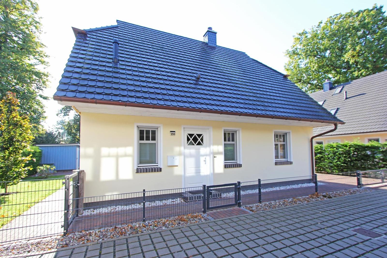 Ein kleines Haus mit gelber Fassade und dunkelblauem Dach, umgeben von Grünflächen und einem Zaun.