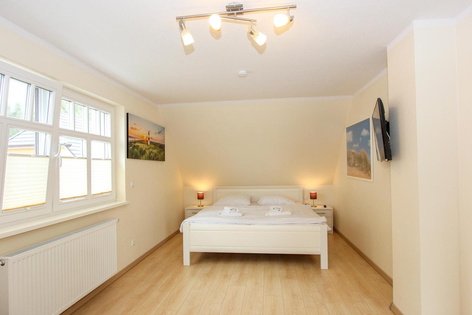 Schlafzimmer mit weißem Bett, Holzboden und Fenster. An den Wänden hängen Bilder und eine Wand-TV.