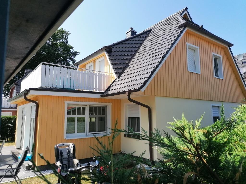 Ein gelbes Haus mit Balkon und Dachgaube. Im Vordergrund sind Grünflächen und ein Fahrrad sichtbar.