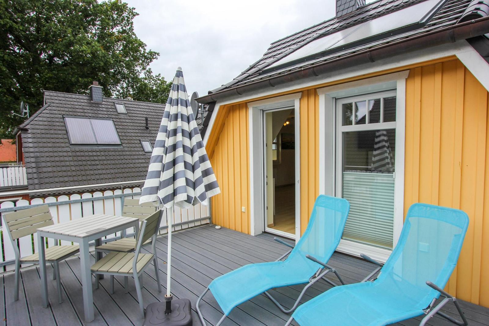 Terrasse mit Tisch, Stühlen und Liegestühlen. Gelb gestrichene Fassade und Glastür.