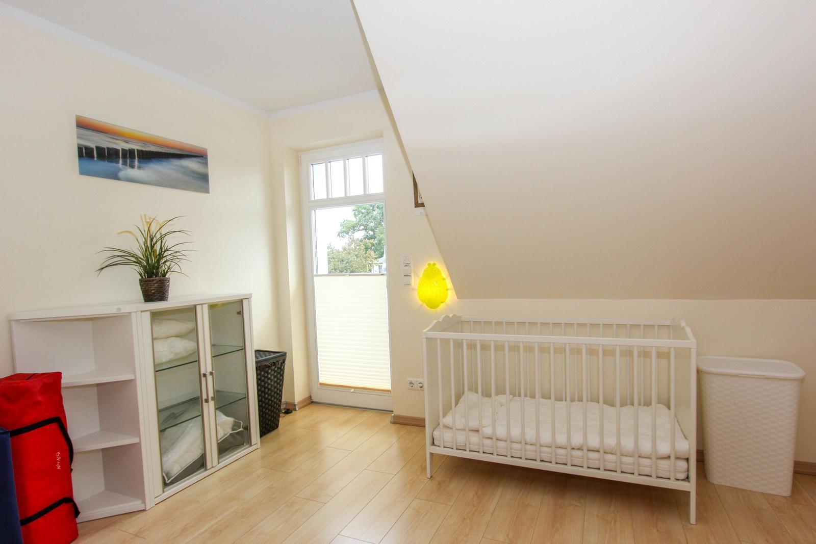 Kinderzimmer mit weißem Kinderbett, Schrank, Pflanze und Fenster.