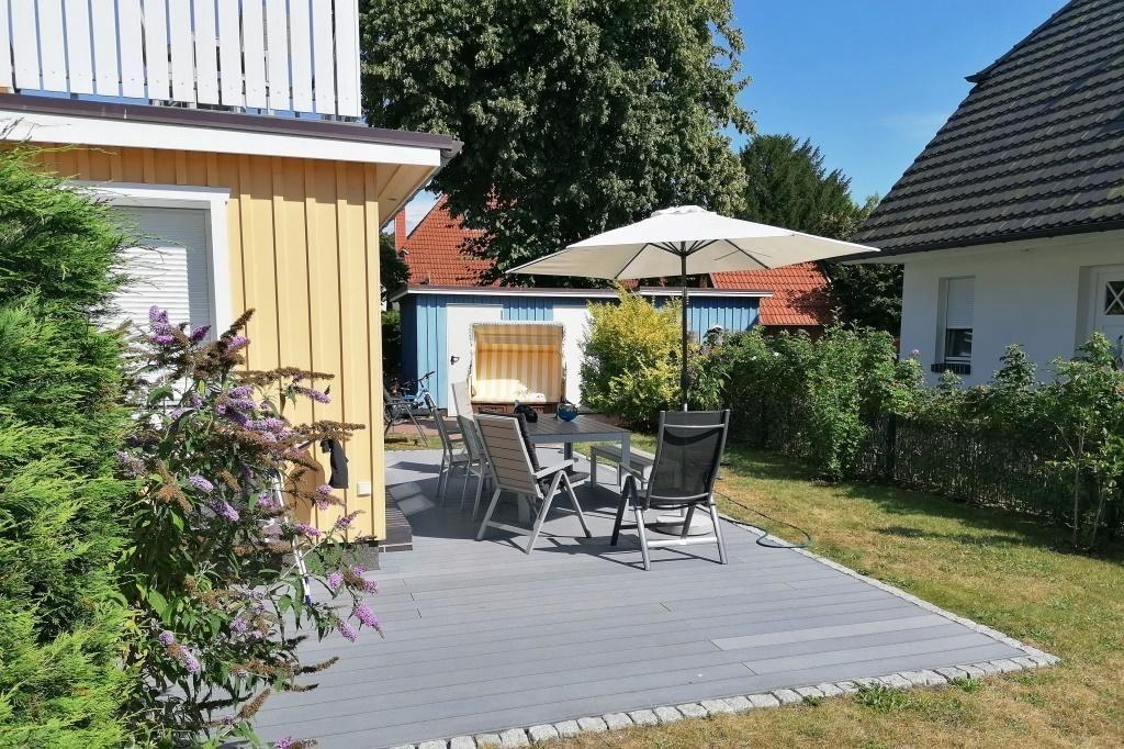 Terrasse mit Tisch, Stühlen und Sonnenschirm. Hintergrund: Garten, Bäume und Nachbarhäuser.