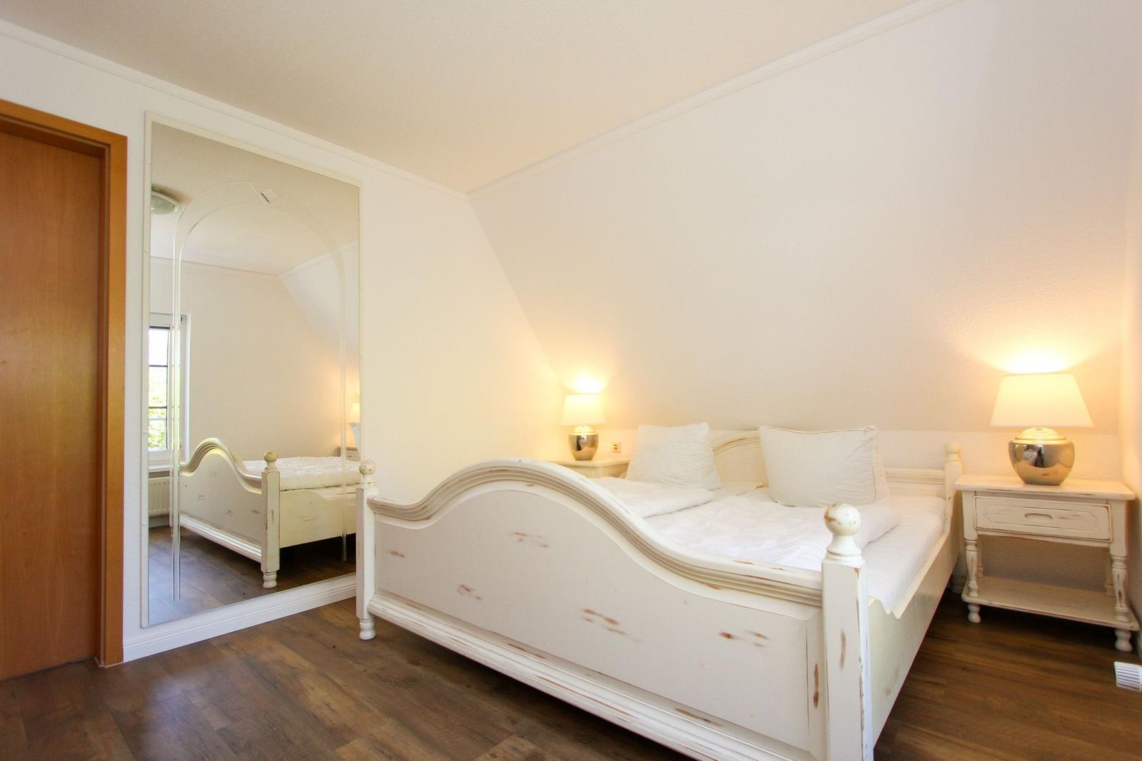 Schlafzimmer mit weißem Bett, Nachttisch und Spiegel an der Wand.