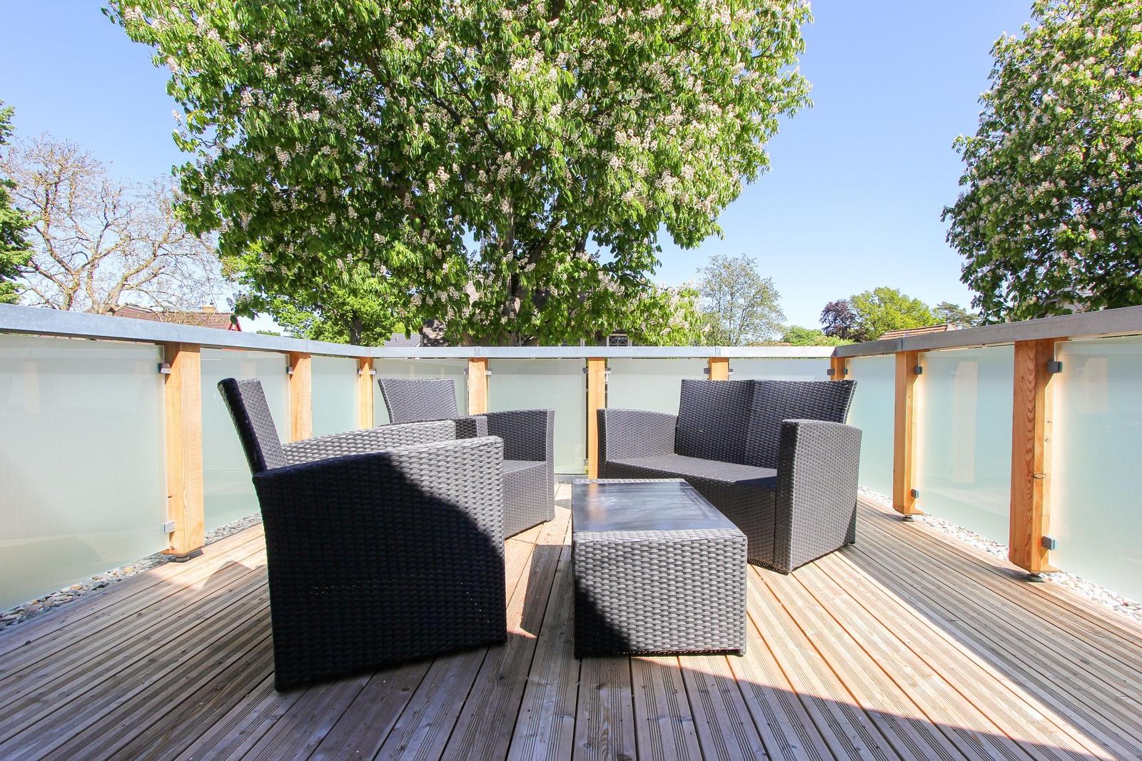 Terrasse mit Gartenmöbeln, Holzdielen und Glasgeländer unter Bäumen.