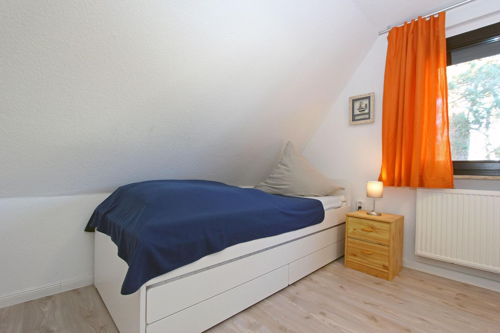 Ein Schlafzimmer mit Dachschrägen, Bett, Nachttisch und Fenster mit orangefarbenen Vorhängen.