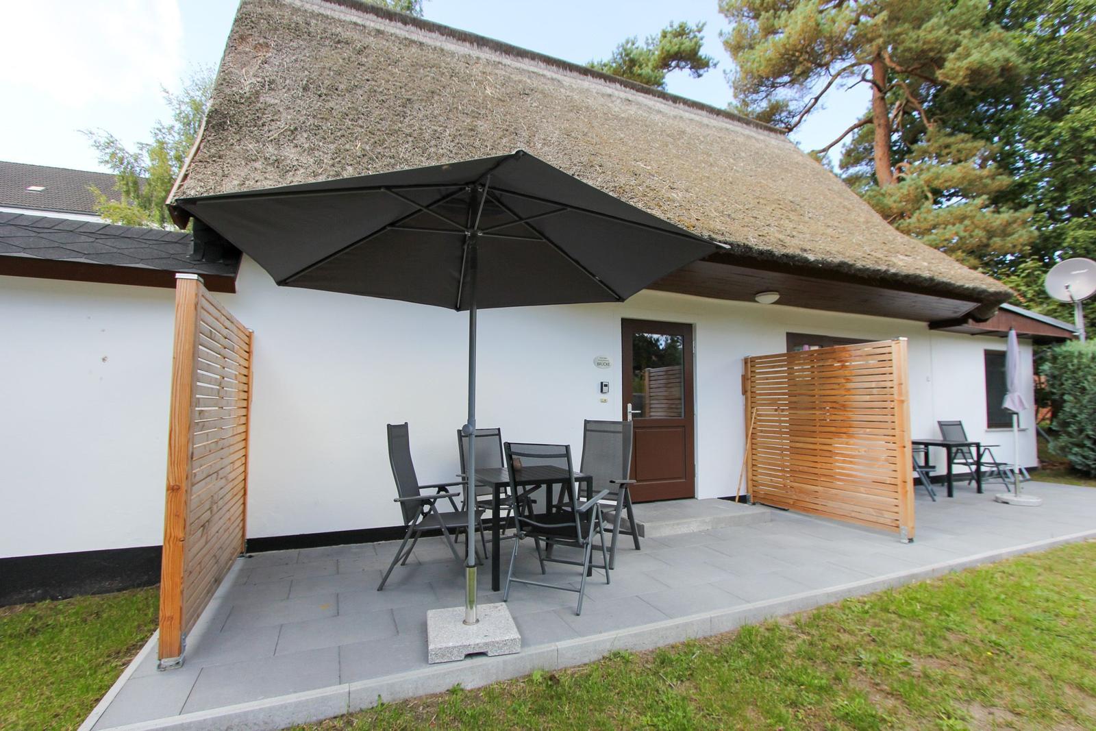 Haus mit Strohdach, Terrasse mit Tisch, Stühlen und Sonnenschirm.