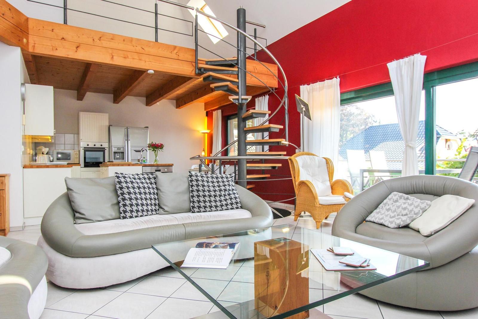 Wohnbereich mit Sofa, Sessel und Glas-Tisch. Hintergrund: Küche und Wendeltreppe.