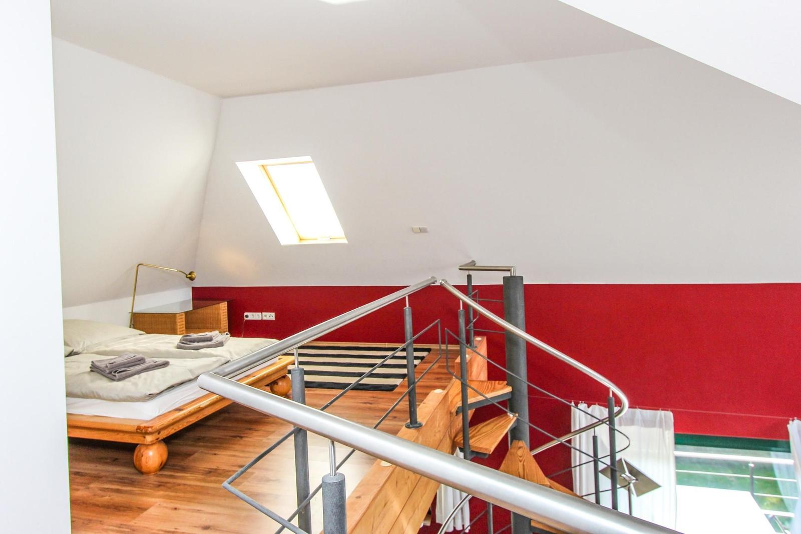 Schlafzimmer mit Dachschrägen, Holzboden und roter Wand. Bett, Treppe und Dachfenster sichtbar.